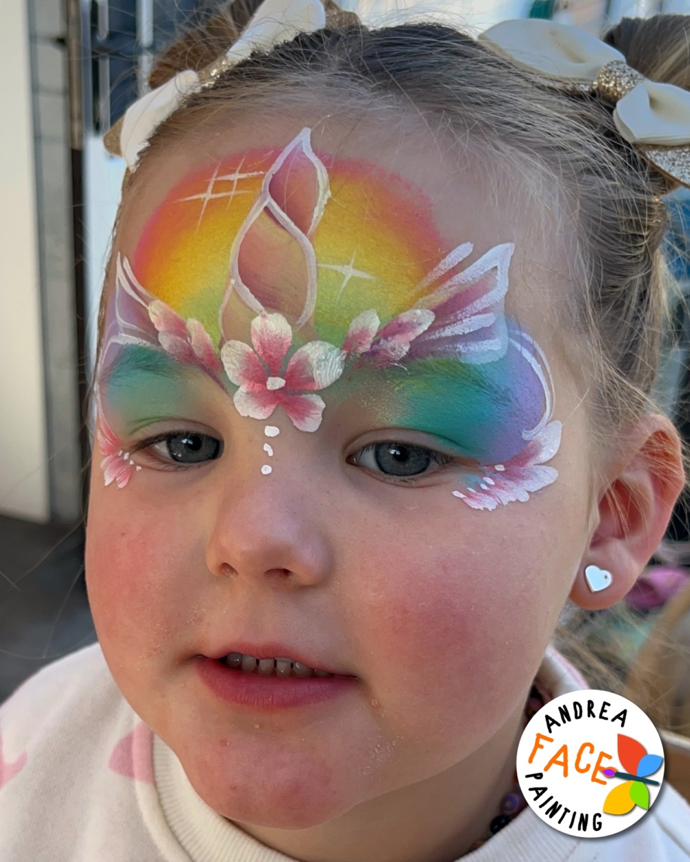 Young girl smiling with a colourful unicorn face paint design featuring rainbow and flowery details